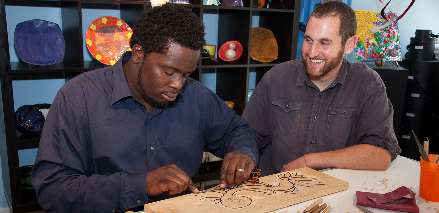 Young man engraving wood while another young man watches the creative process